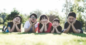 Mindfulness and Parenting , A happy family of five lying on the grass outdoors, resting their faces on their hands in a playful and relaxed manner, enjoying a mindful moment together in nature.